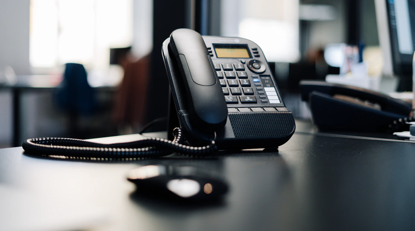 Black IP desk phone on an office desk with blurred workspace behind