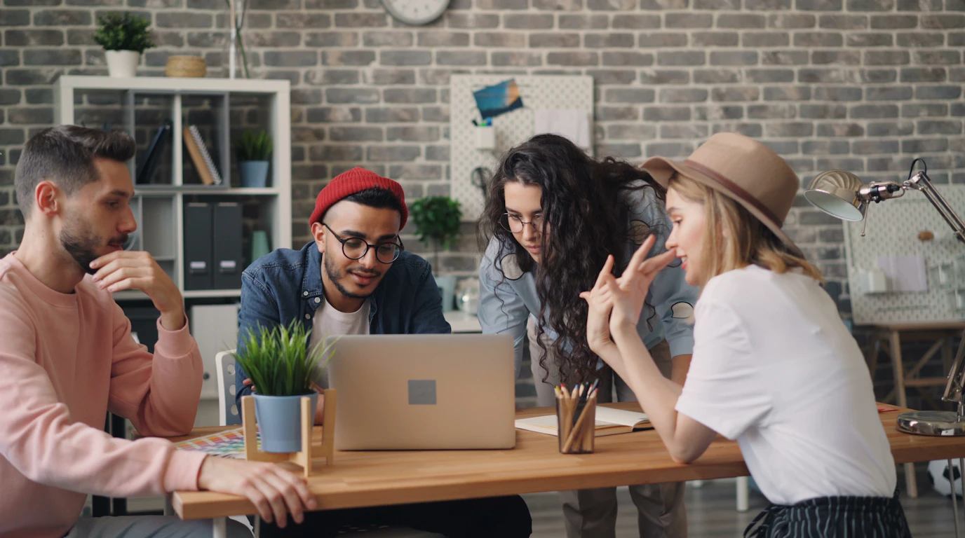 Small team gathered around a laptop at a wooden table in a brick-walled office